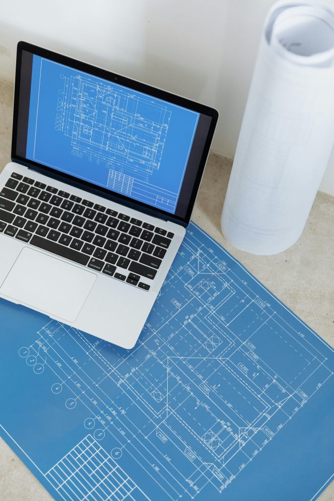 pexels-photo-8470810-8470810 Laptop showing engineering blueprint with rolled paper and drawings on table.